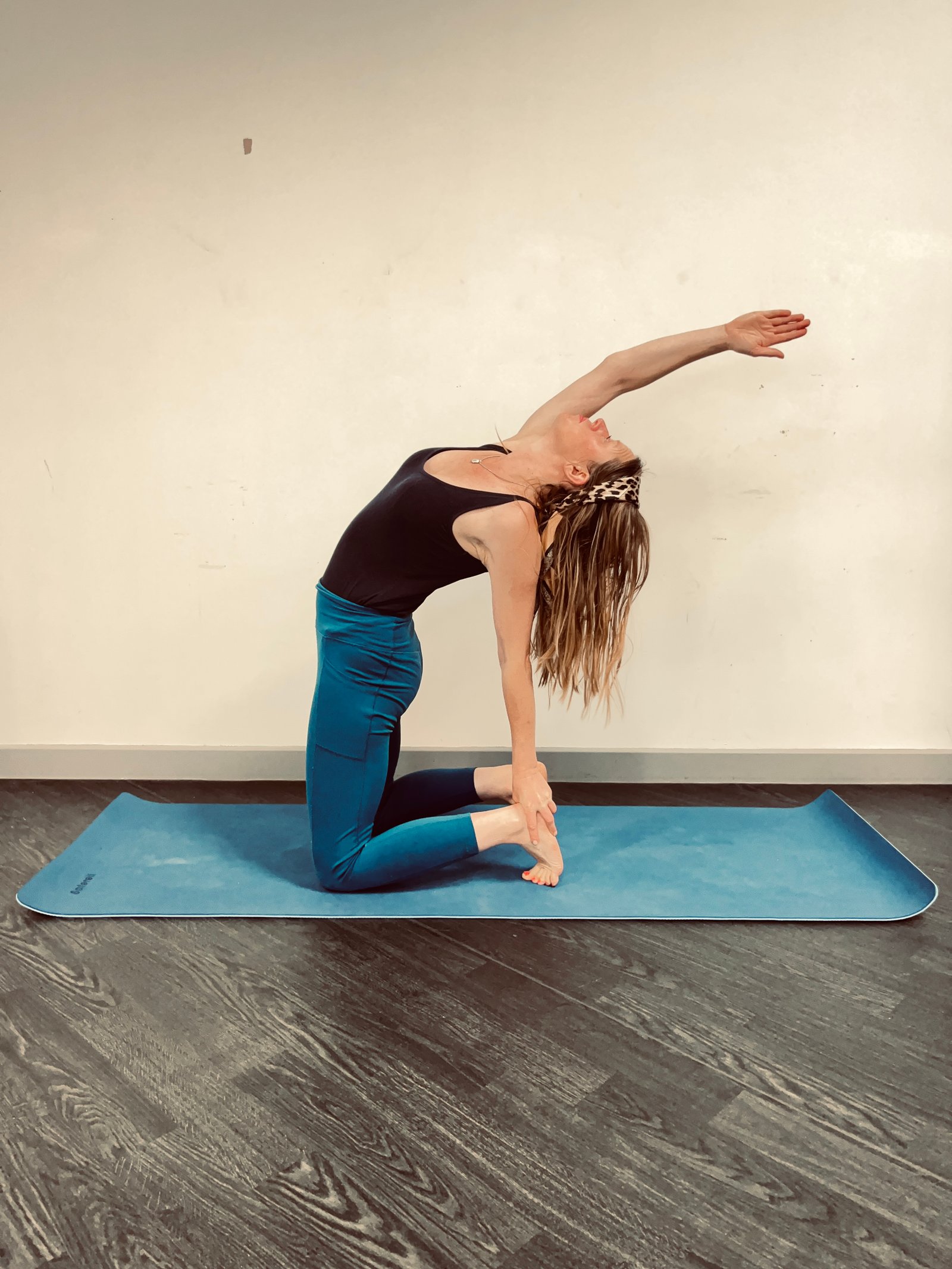 Abi kneeling in a supported backbend on a blue mat, one arm sweeping overhead.