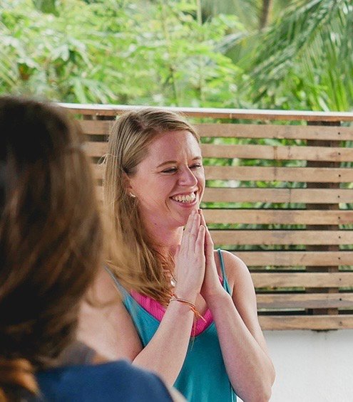 Abi laughing, hands together at her chest, on a covered terrace surrounded by greenery.